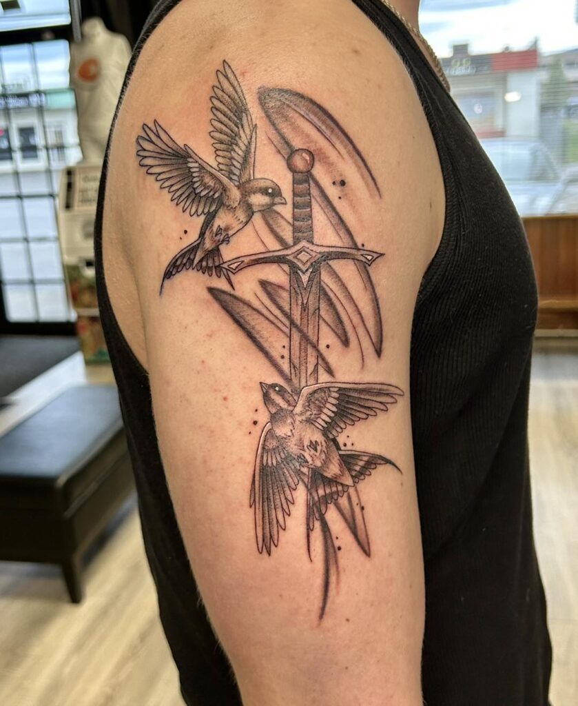 Fine line bird and sword tattoo by Akira featuring two swallows with soft black and grey shading and detailed wings on upper arm.