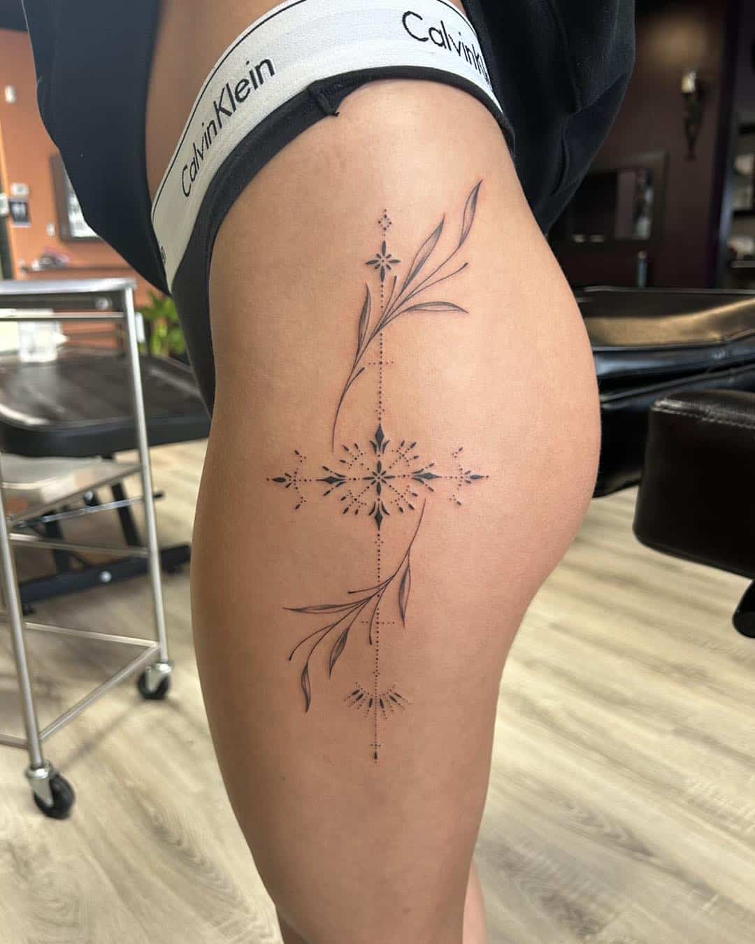 Fine line ornamental side thigh tattoo by Akira featuring geometric starburst design with delicate botanical elements and minimal black ink detailing.