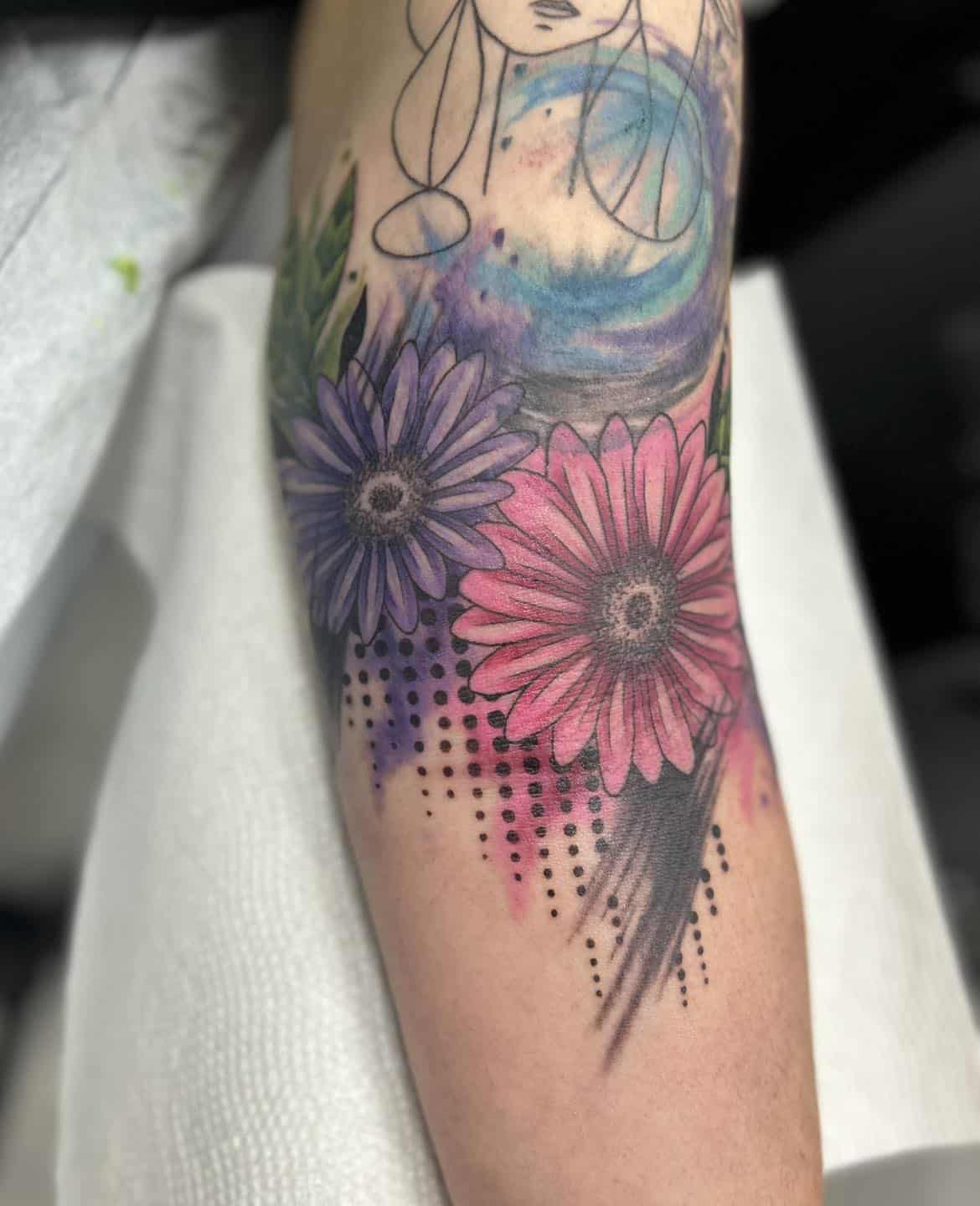 Watercolor floral tattoo by Akira featuring pink and purple gerbera daisies with abstract color splashes and dotwork detailing on forearm.